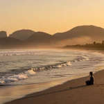 Natureza e Bem-Estar: Conectando-se em 2025 para uma Vida Plena Pessoa meditando em uma praia brasileira ao nascer do sol, com ondas suaves e vegetação exuberante ao fundo, transmitindo paz.
