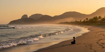 Pessoa meditando em uma praia brasileira ao nascer do sol, com ondas suaves e vegetação exuberante ao fundo, transmitindo paz.